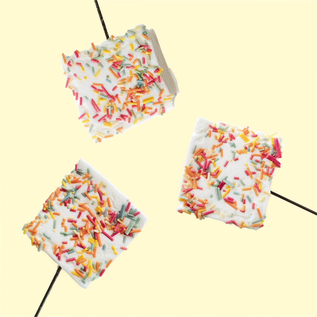 Three large square birthday cake marshmallows with rainbow sprinkles on a light yellow background