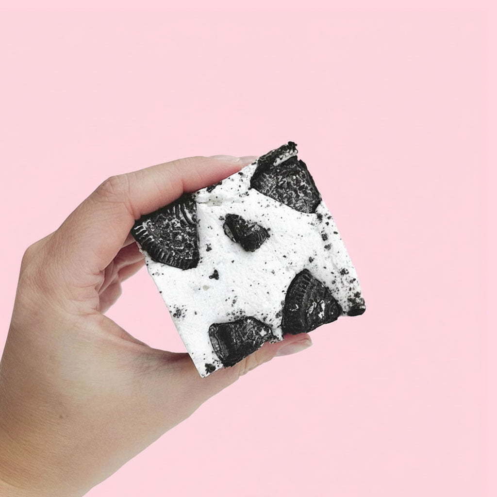 Hand holding a large gourmet marshmallow with Oreo cookie pieces against a pink background